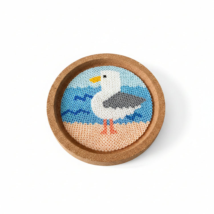 A round cork coaster featuring a handcrafted bead art design of a seagull in light blue, white, gray, yellow, and orange, displayed on a wooden surface.